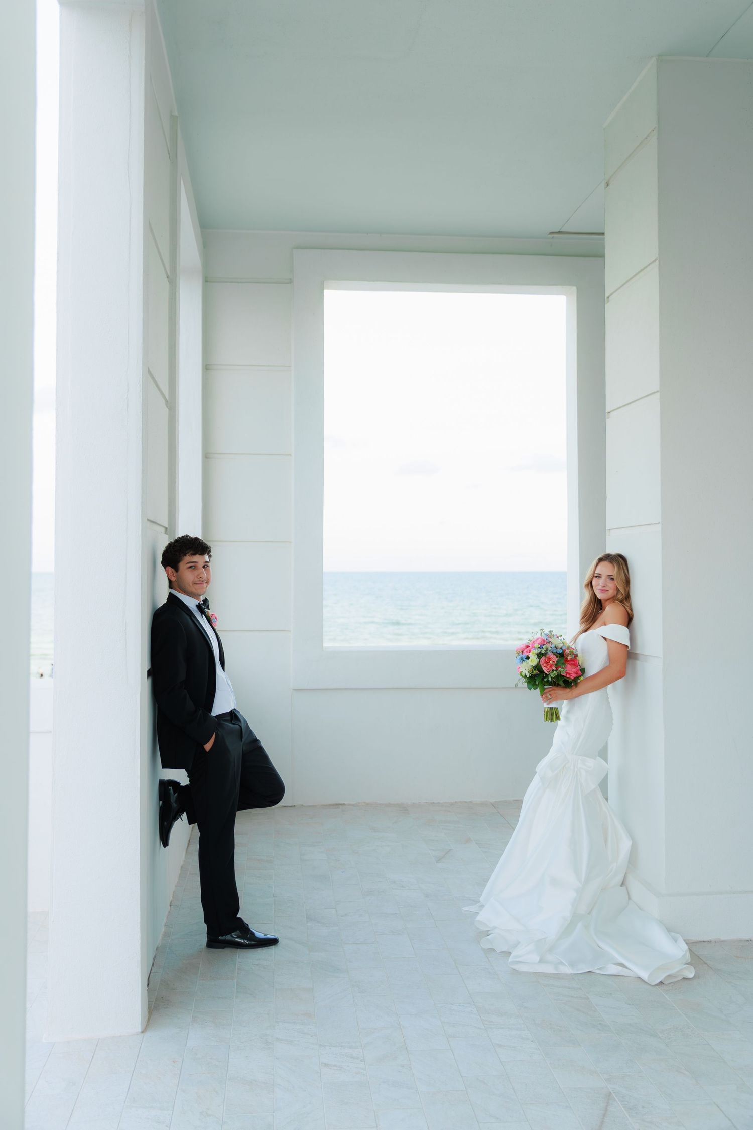 A couple in wedding attire poses in an elegant white archway overlooking the ocean on their special day.