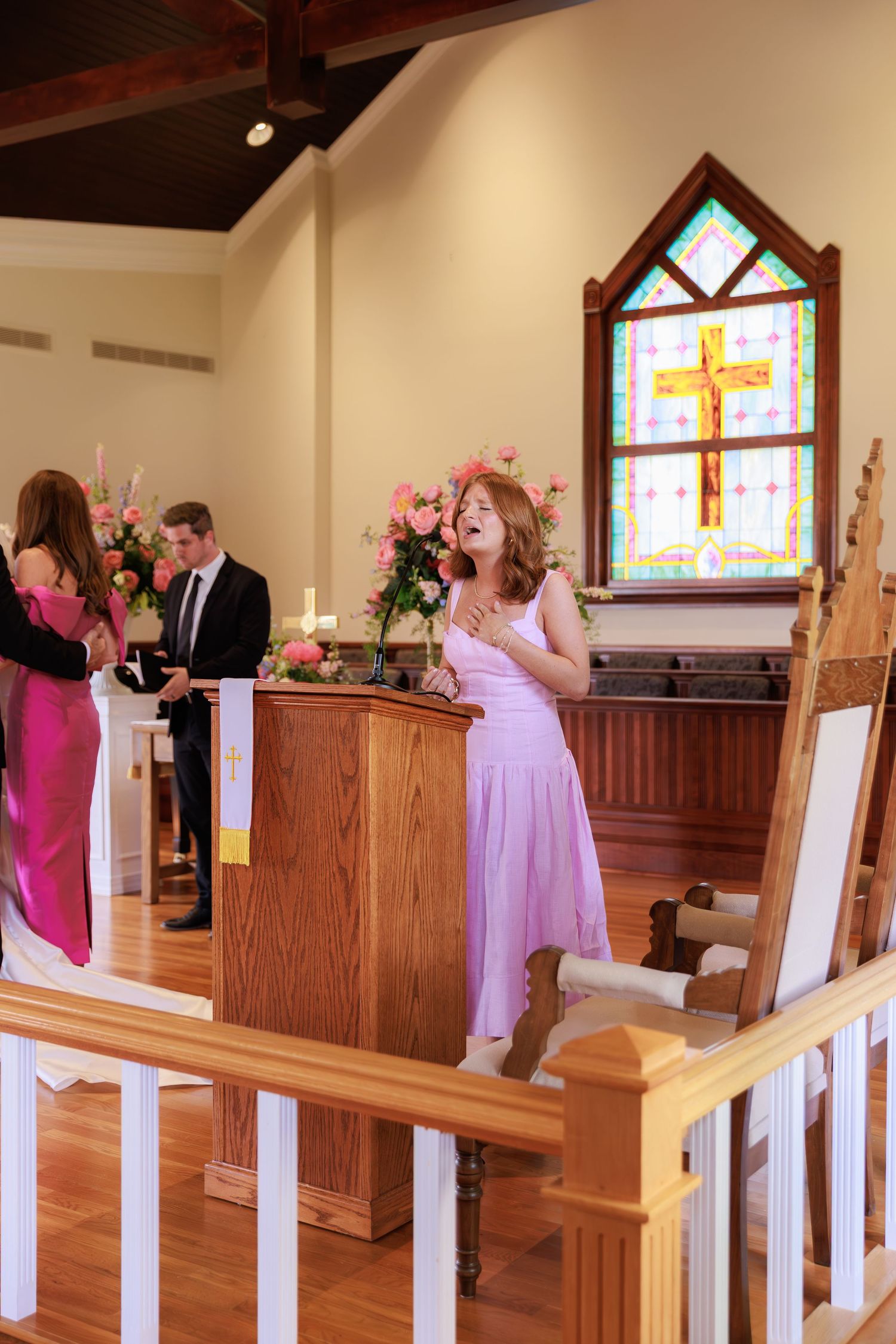 People in formal attire gather near a podium in a chapel with a stained glass window featuring a cross.