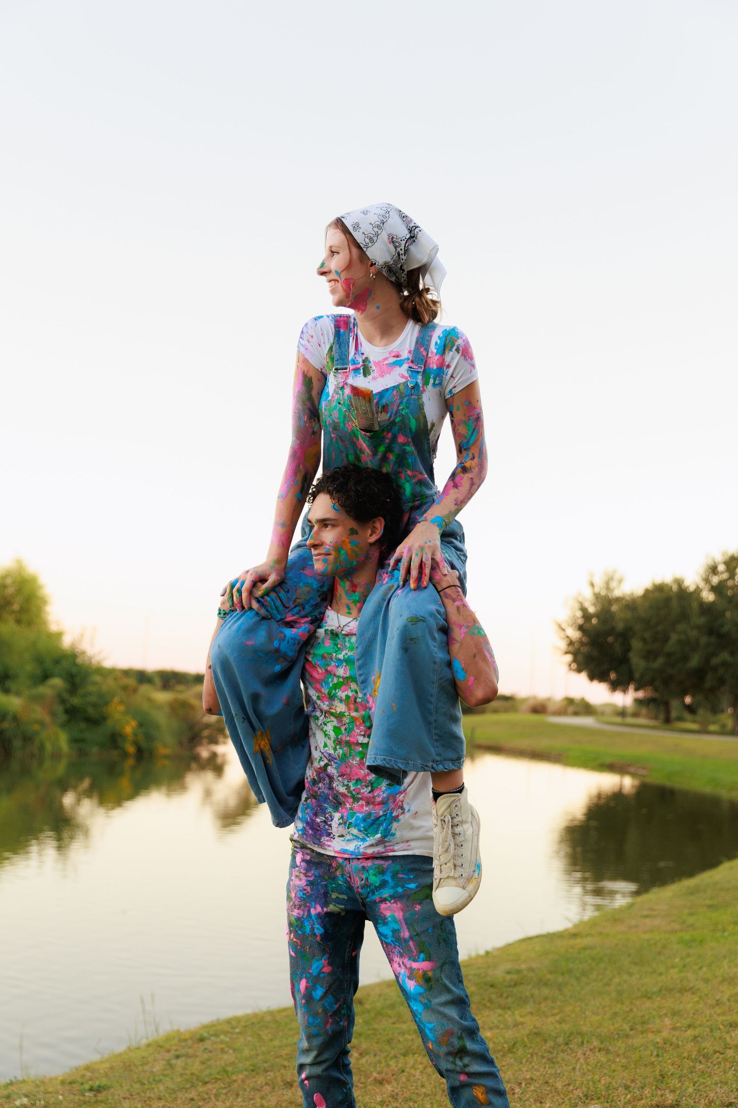 Two people in matching floral outfits pose playfully by a lake at sunset with one sitting on the other's shoulders.