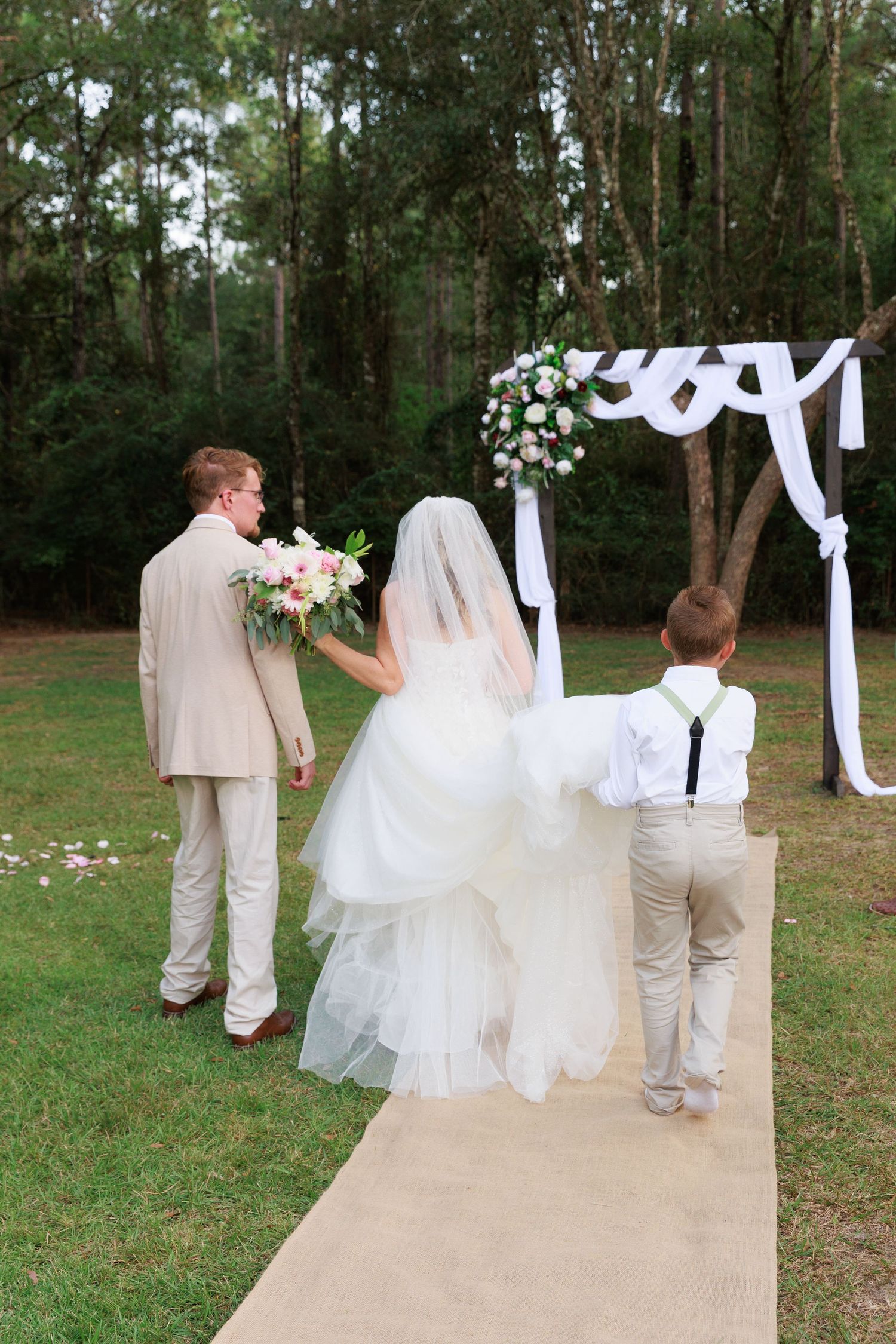 Wedding ceremony setup with tan aisle runner and white draped arch in a serene wooded outdoor setting.