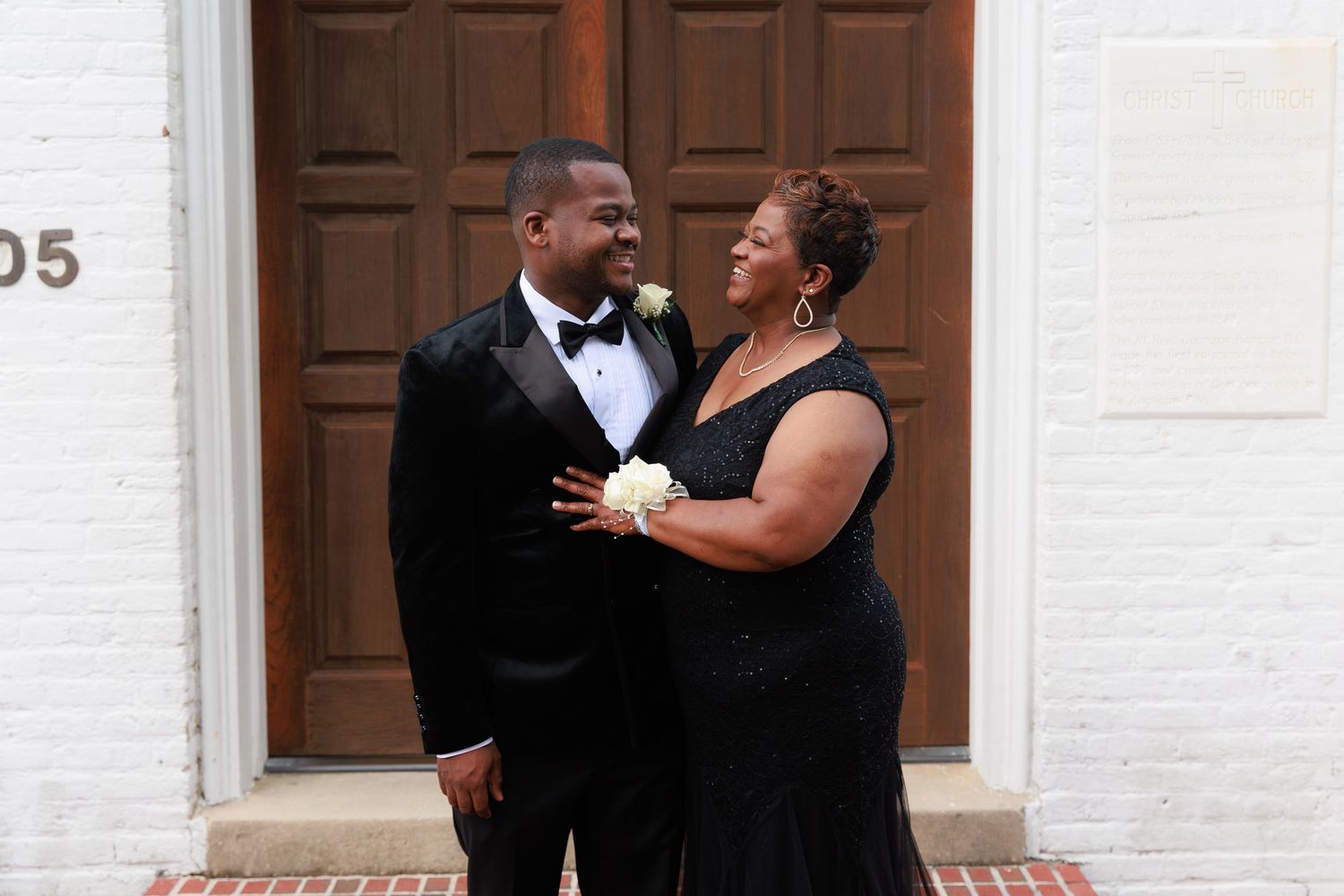 Wedding party members in formal black attire share a joyful moment outside a white building with wooden doors.