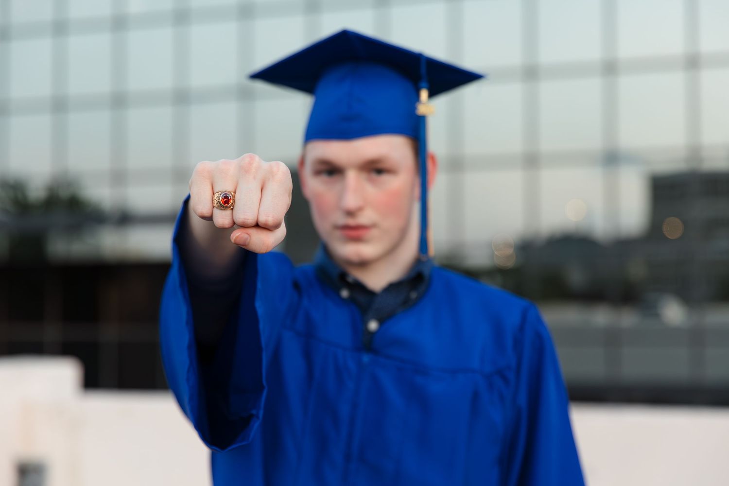A graduate in blue cap and gown proudly displays a class ring toward the camera outdoors.
