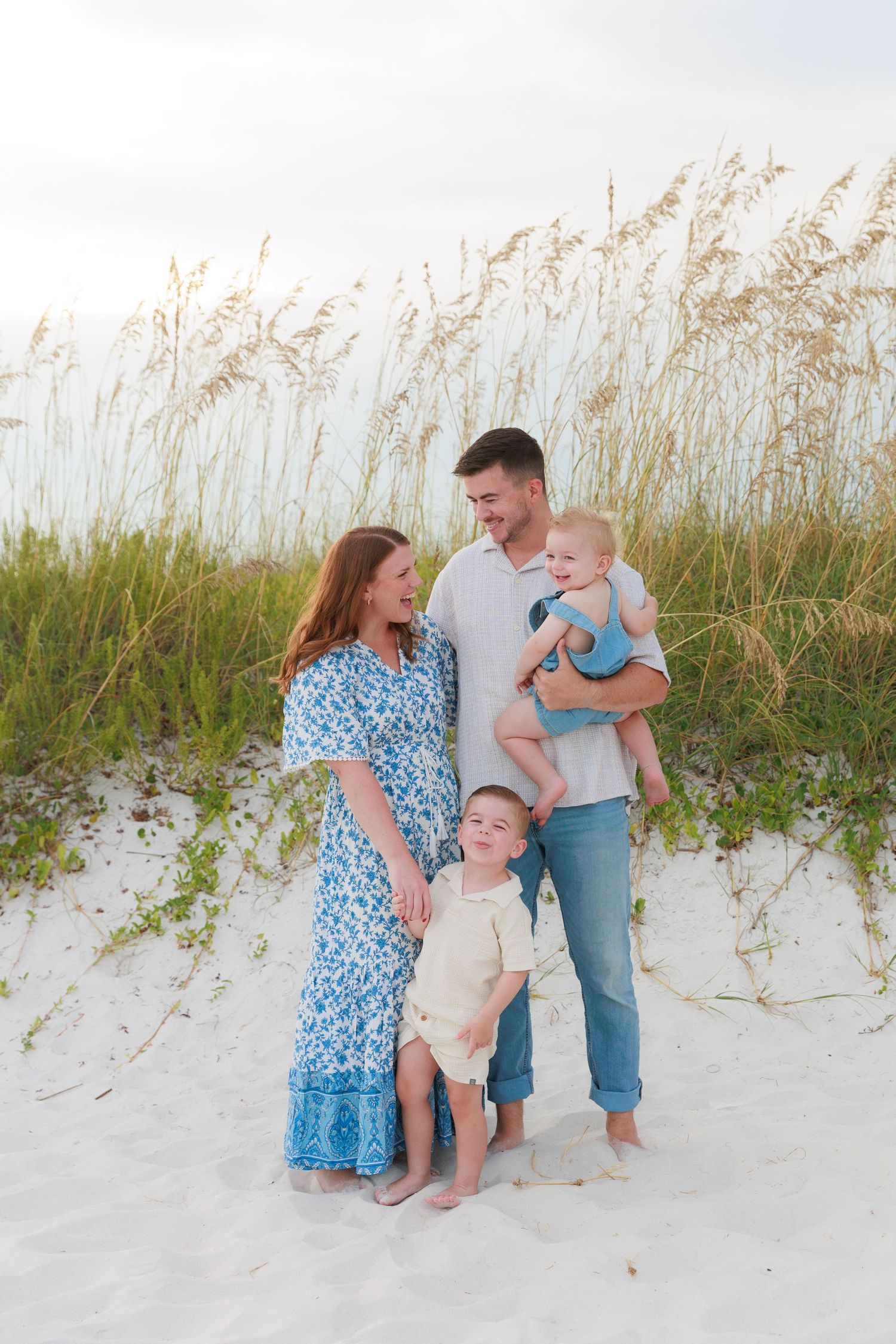 Happy family of four poses on white sand beach dunes, mother in blue floral dress, father holding baby in blue romper.