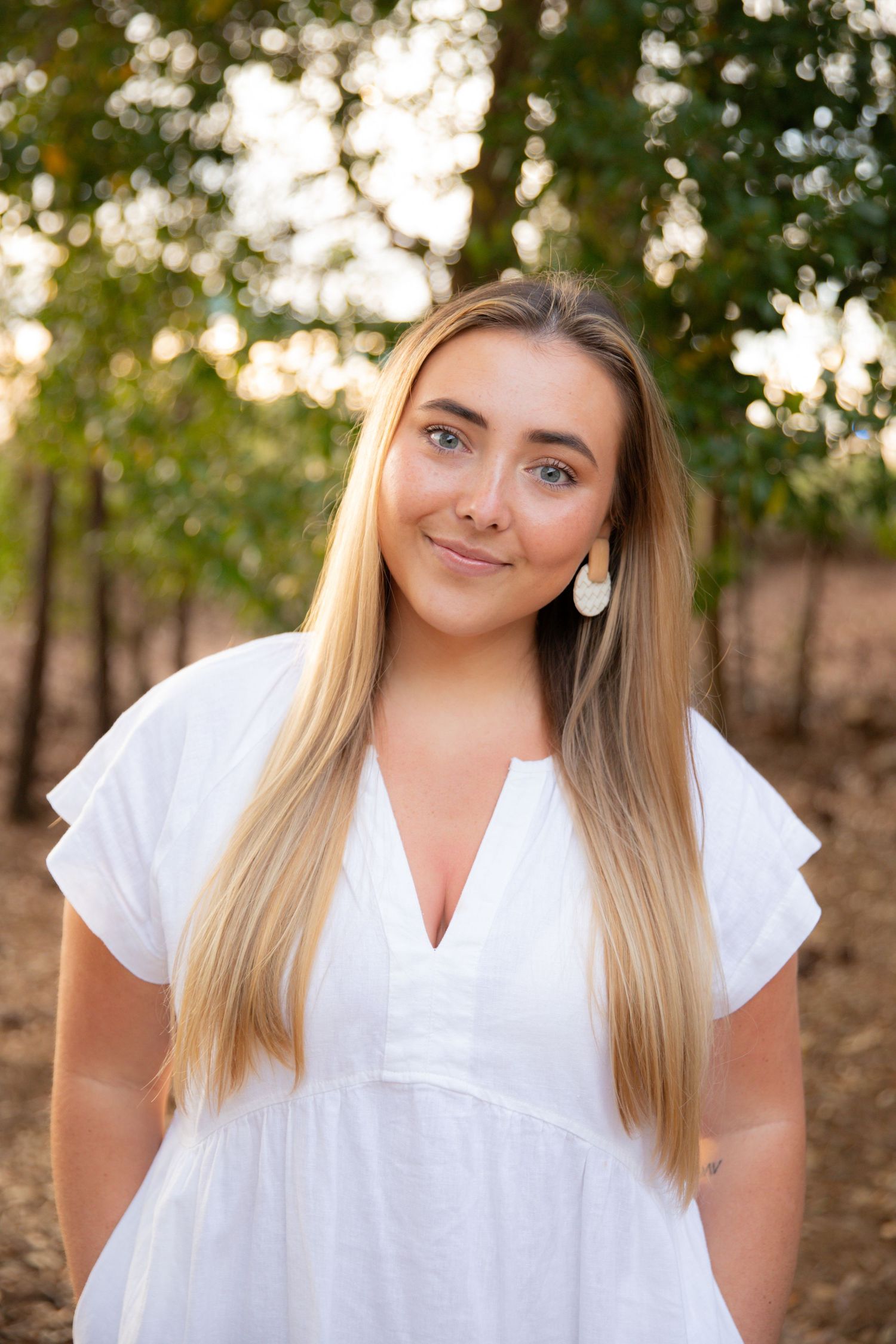 Young woman with long blonde hair wearing a white dress and earrings smiling outdoors among green trees.