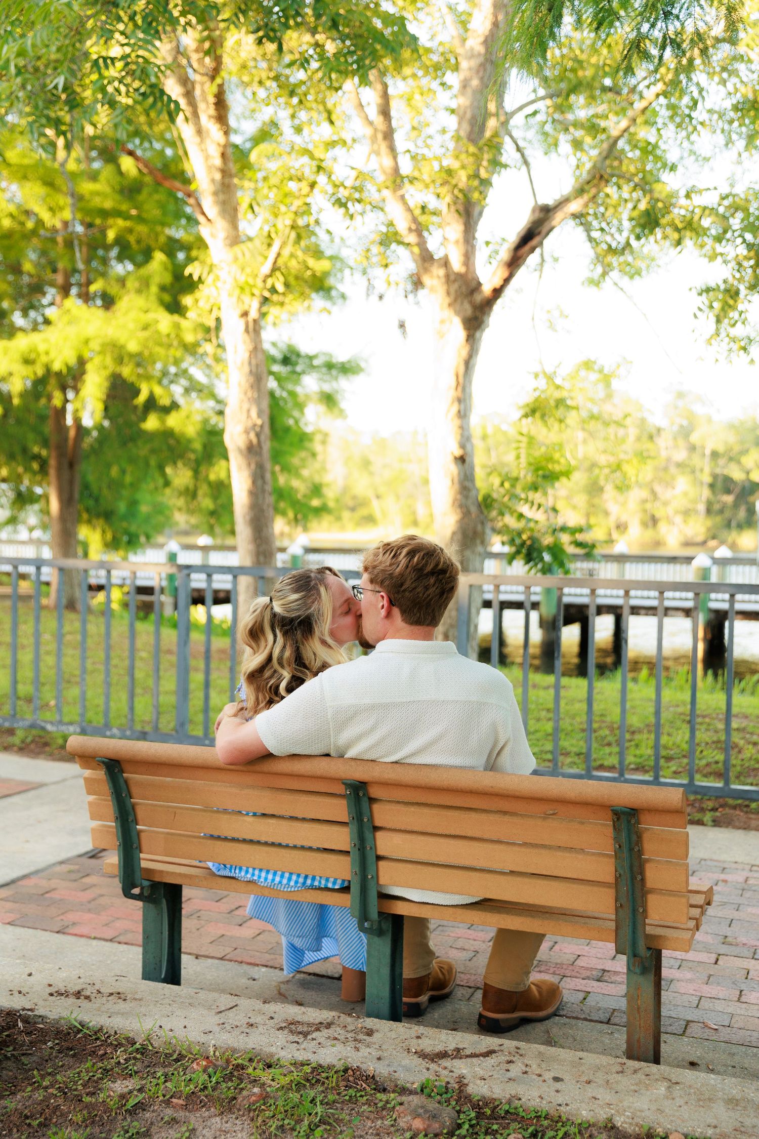 Couple sharing a romantic kiss on a wooden park bench surrounded by lush green trees near a waterfront.