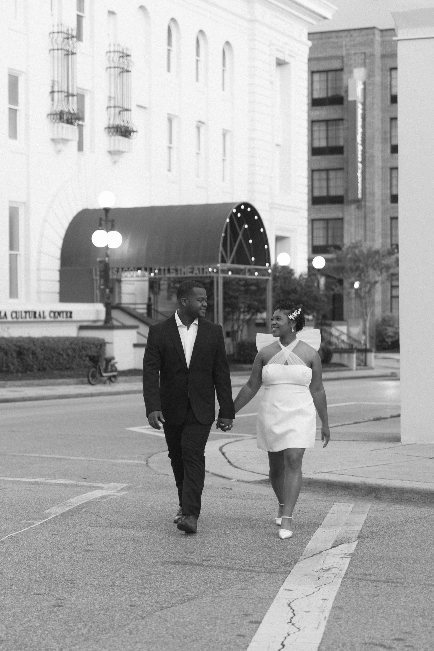 Black and white photo of a couple holding hands walking on a city sidewalk, dressed in wedding attire.