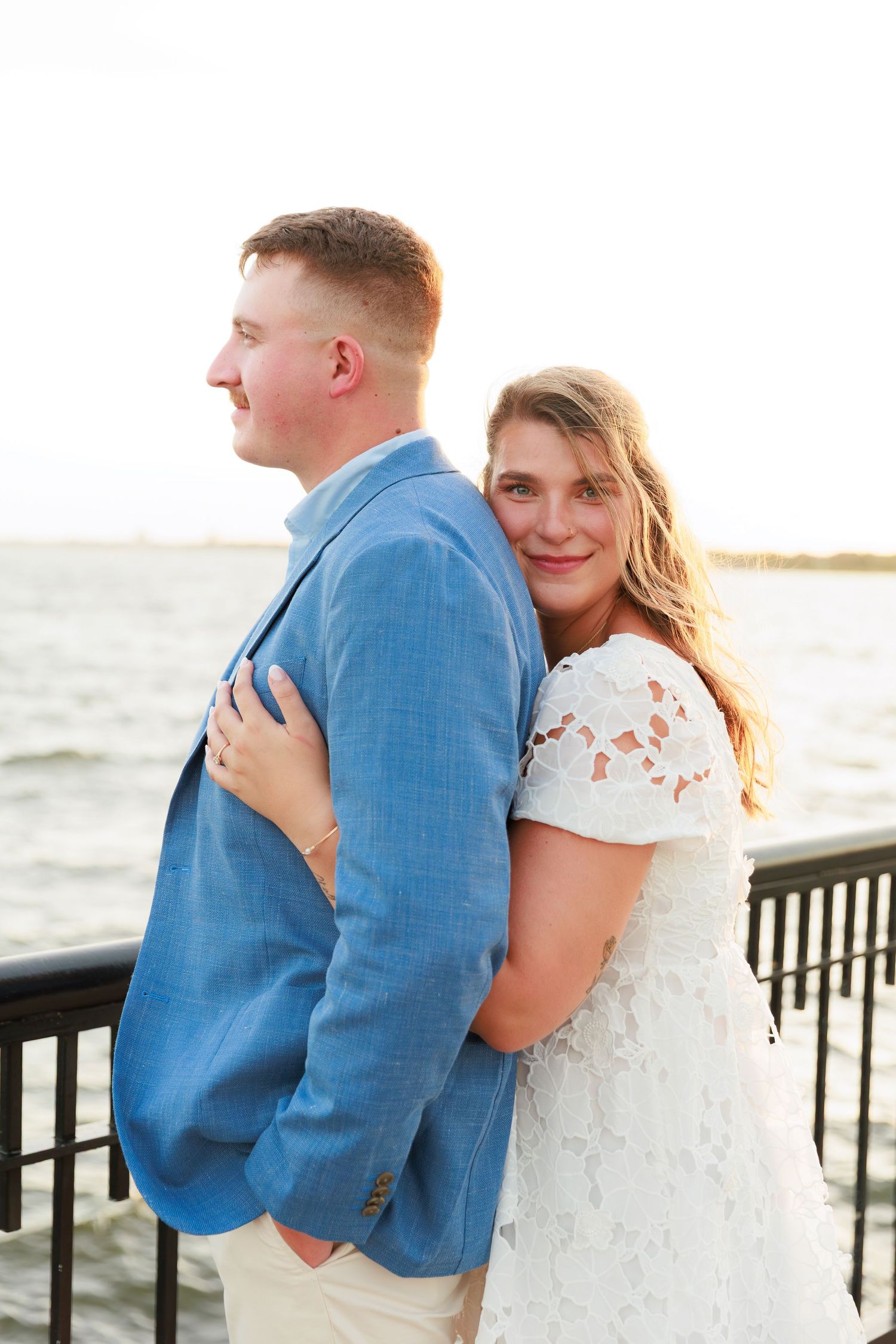 Woman in white lace dress hugging man in blue blazer from behind near a waterfront railing at sunset.