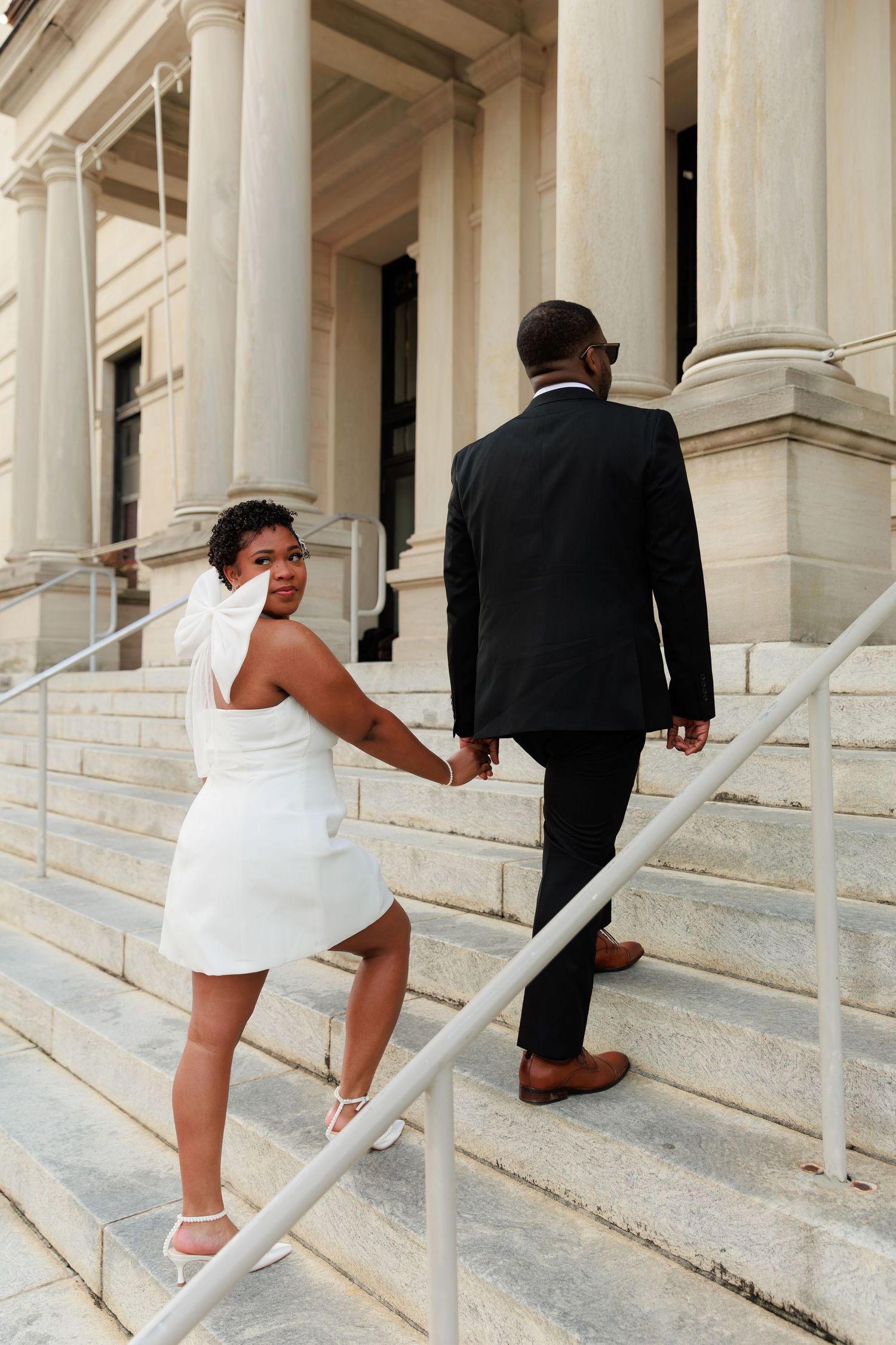 Couple in wedding attire climbing stone steps of a grand building, bride in white mini dress looks back smiling.