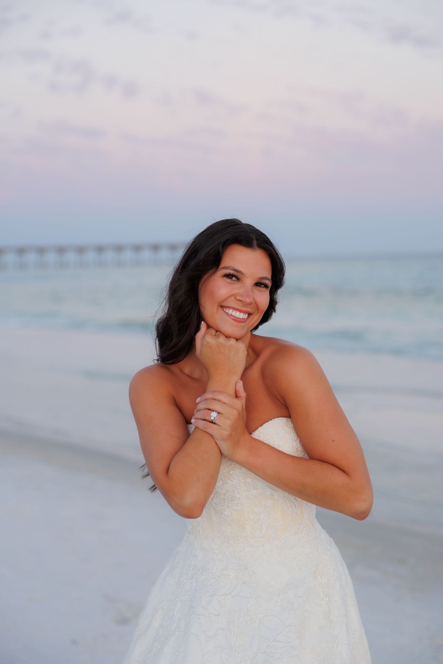 Radiant bride in strapless white lace gown smiling on beach at dusk, showcasing engagement ring near ocean pier.