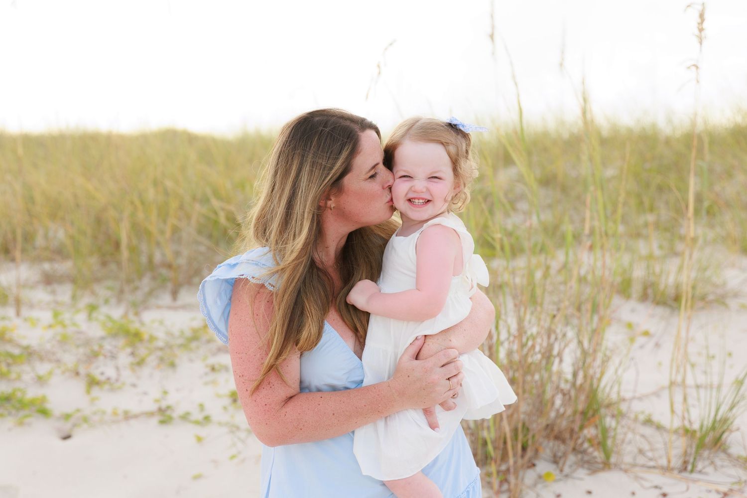 A mother in a blue dress kisses her toddler daughter in white on a sandy beach with sea grass.