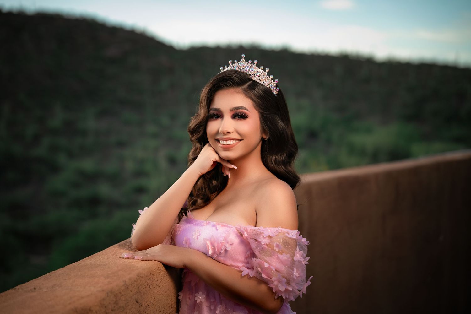 A smiling person in a pink off-shoulder gown and crown poses outdoors against a scenic mountain backdrop.