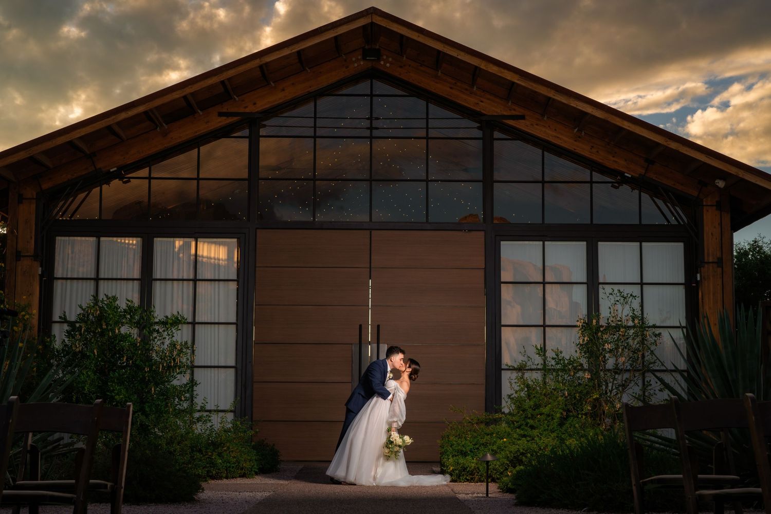 Silhouetted couple pose dramatically against modern glass building facade at sunset.
