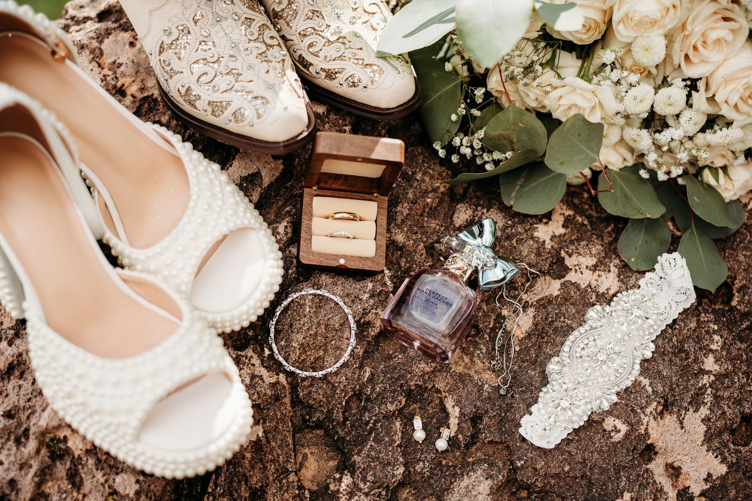 Wedding accessories including lace shoes, perfume bottle, rings and white floral bouquet arranged on rustic surface.