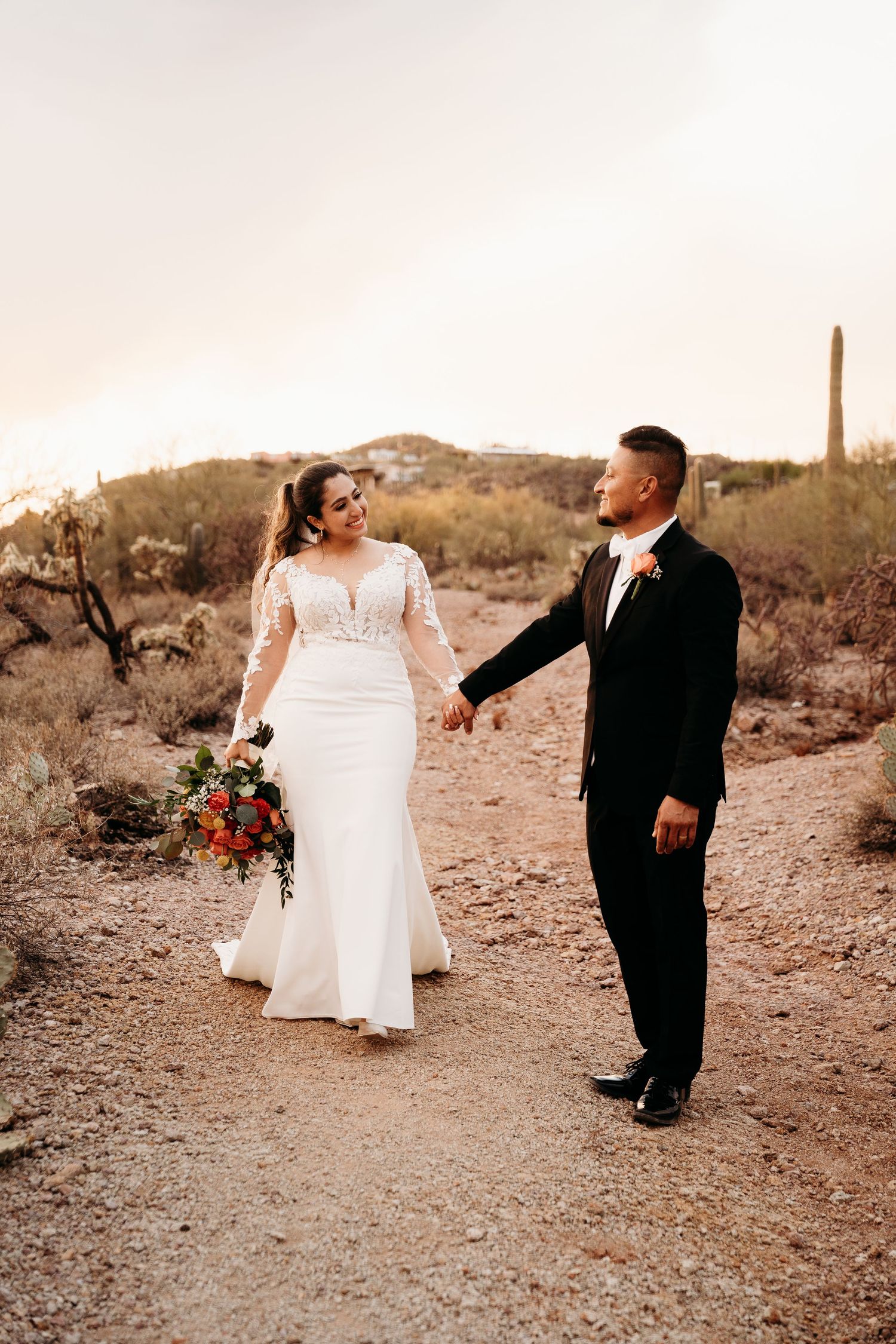 A bride in a lace-sleeved gown and groom in tuxedo walk along a desert trail at sunset with saguaro cacti in background.