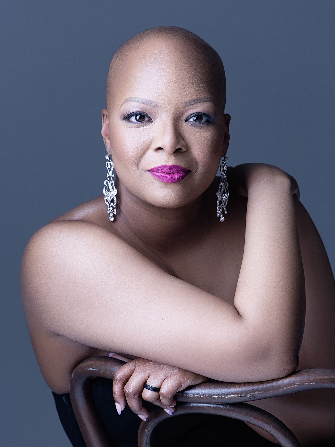 Elegant woman with shaved head wearing chandelier earrings and bold pink lipstick poses confidently in a studio portrait.