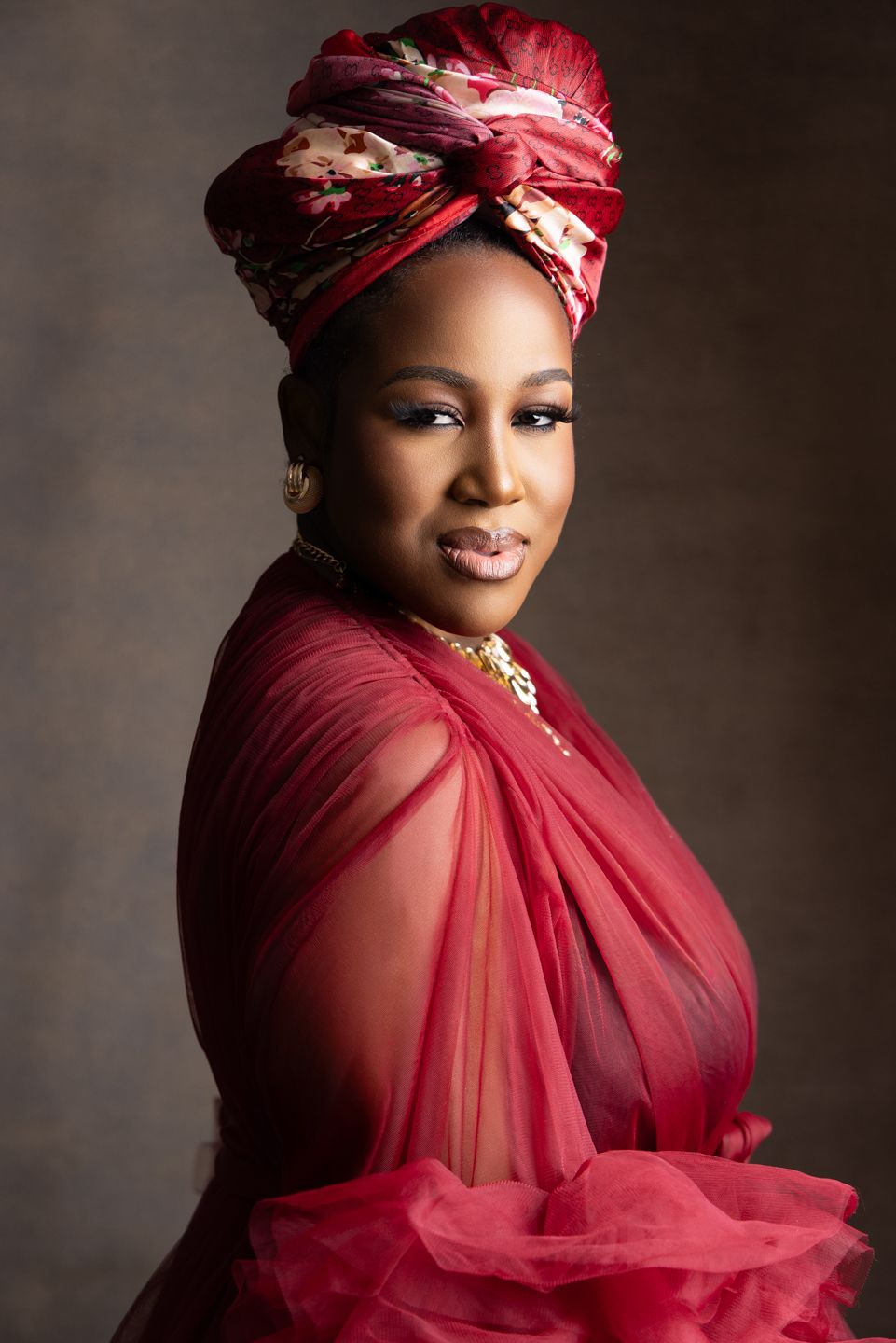 Woman in elegant red tulle dress and matching floral head wrap poses gracefully in a professional portrait.