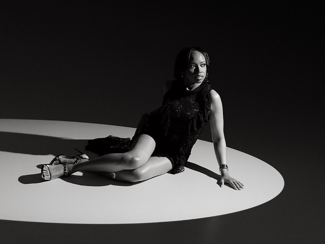 Black and white photo of a woman in a black dress posing elegantly on a circular lit floor in a dark studio.