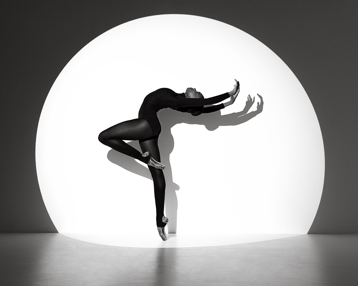 Silhouetted dancer performs graceful movements against a bright white circular backdrop in black and white.