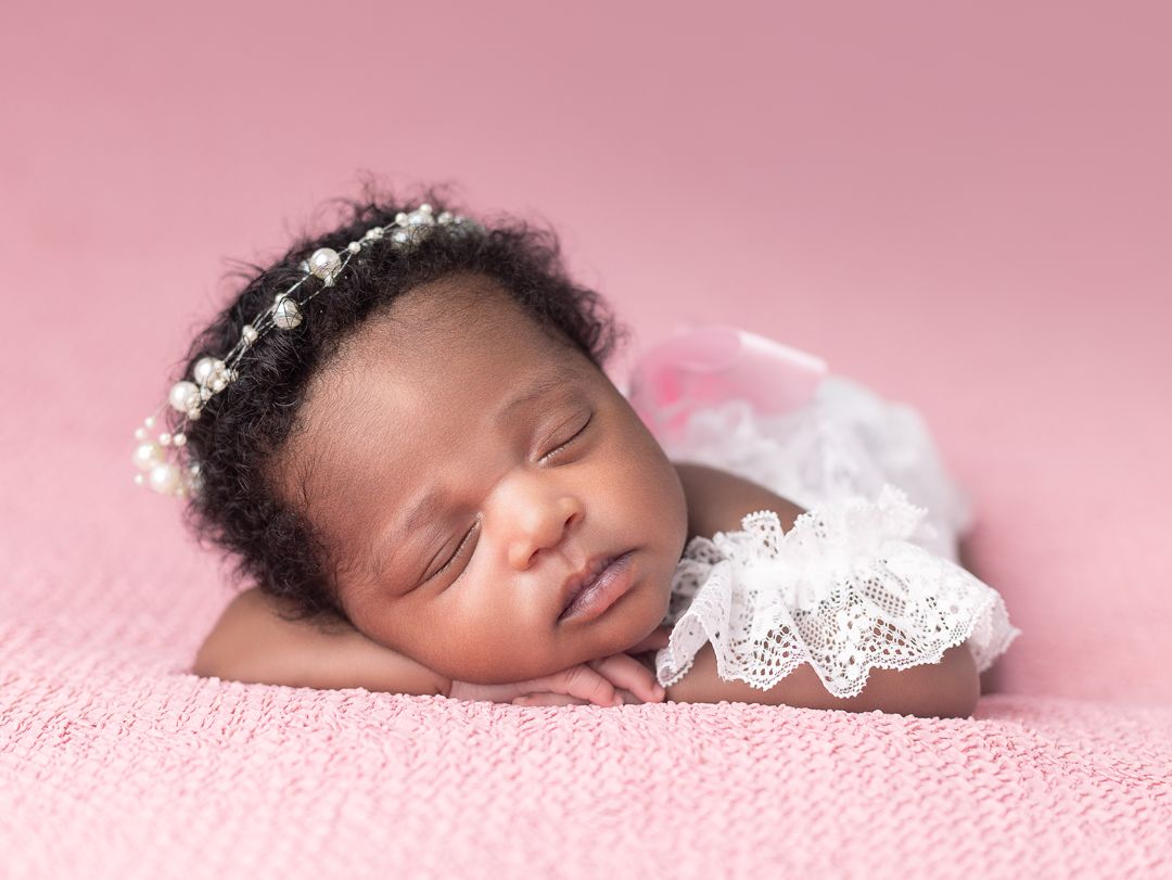 Peaceful newborn baby girl sleeping on pink blanket wearing white lace outfit and floral pearl headband.