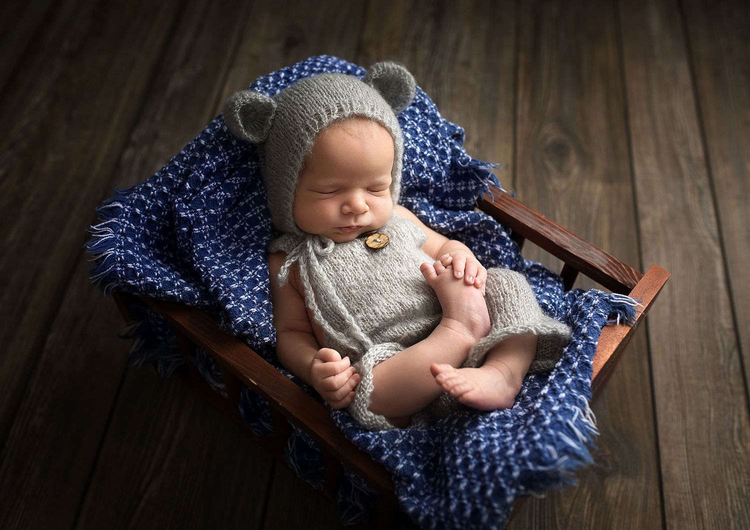 Sleeping newborn baby in gray knit bear bonnet and romper resting on blue blanket in wooden crib.