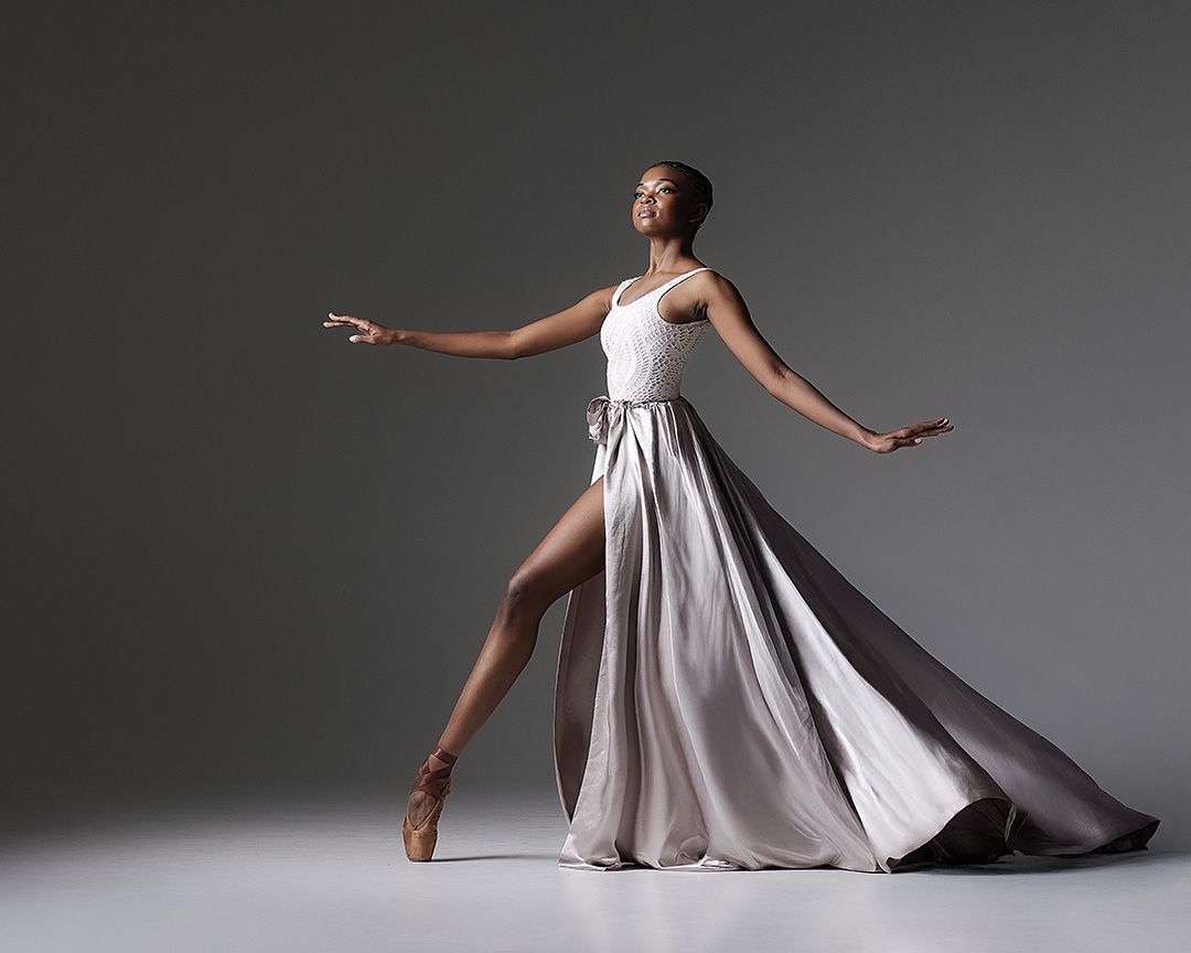 A ballet dancer in a flowing silver gown strikes an elegant pose with arms outstretched against a dark background.