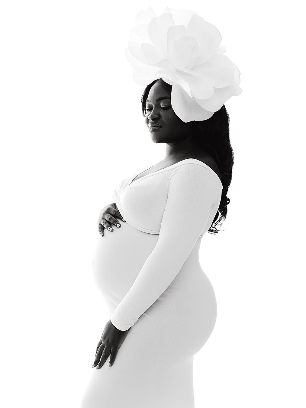 Black and white maternity photo of a pregnant woman in a white dress and large floral headpiece, cradling her baby bump.