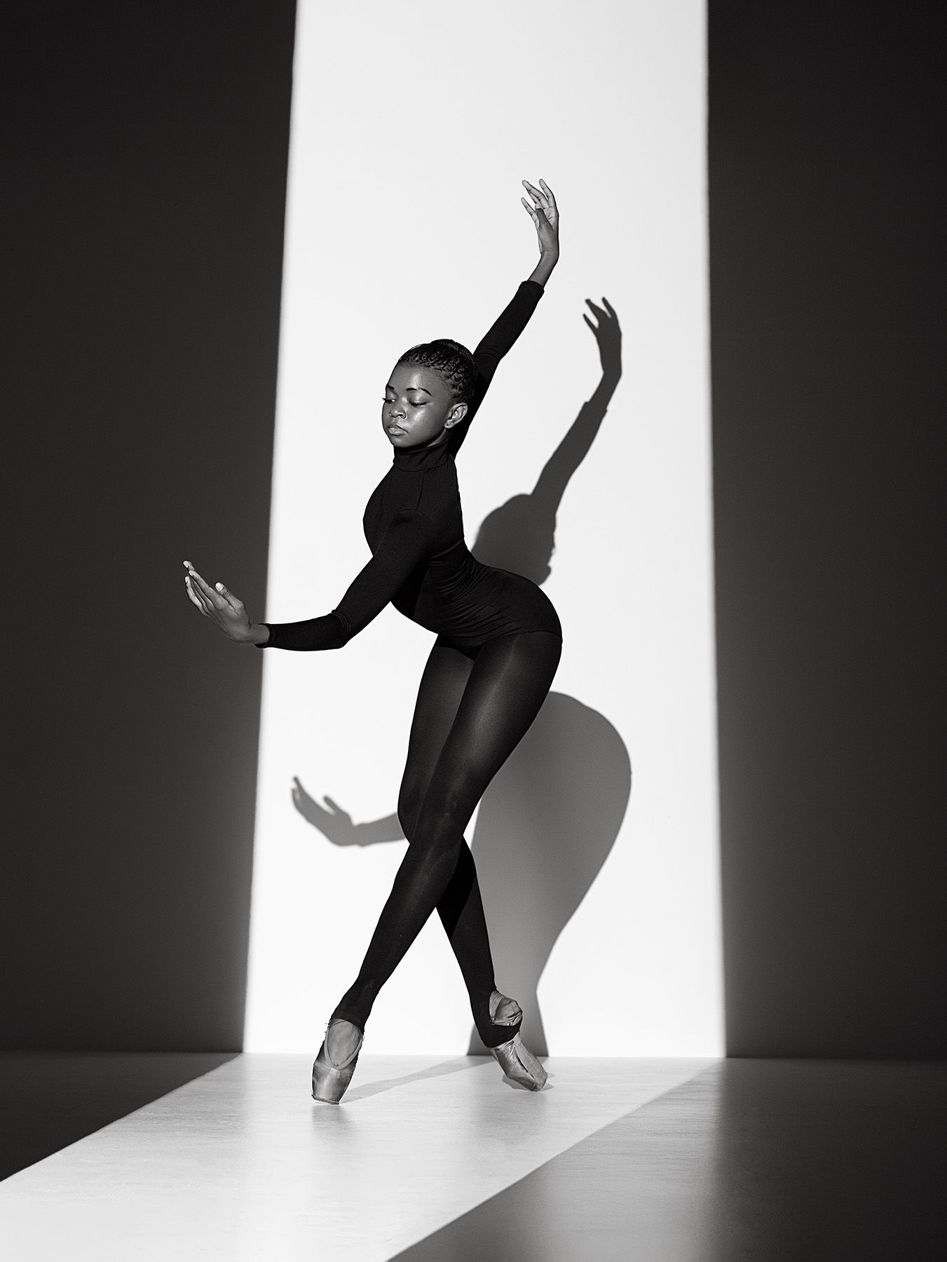 Graceful ballet dancer in black leotard performs elegant pose against dramatic light and shadow background.