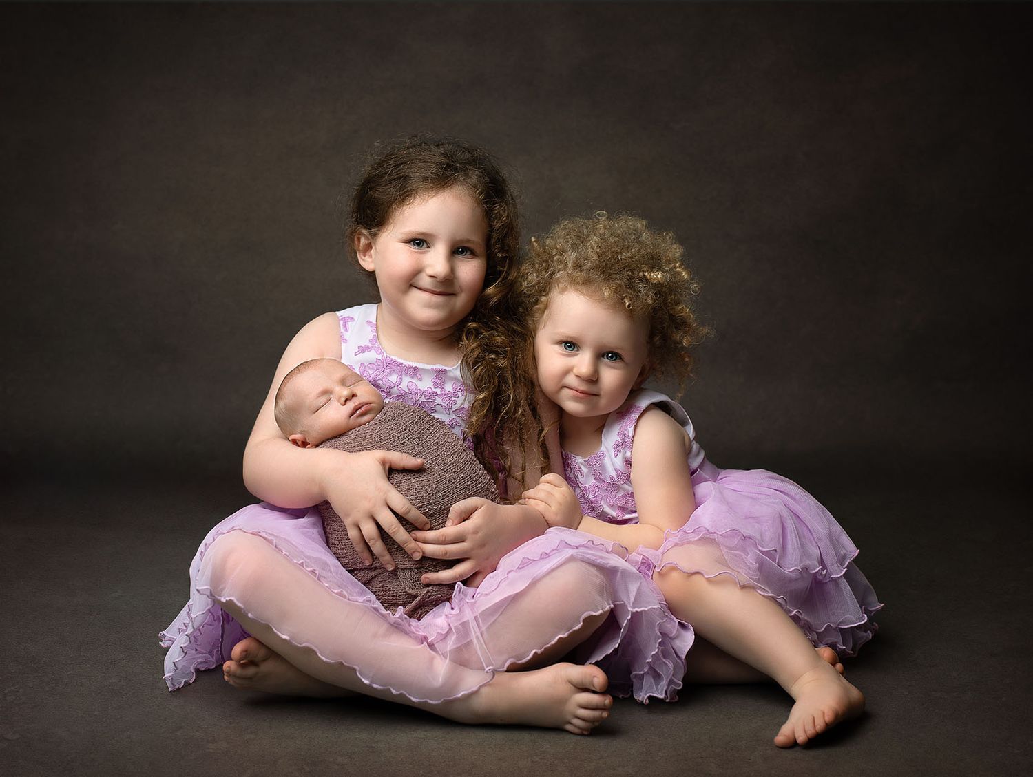 Two young girls in purple dresses pose with a sleeping newborn baby in a professional portrait photo.