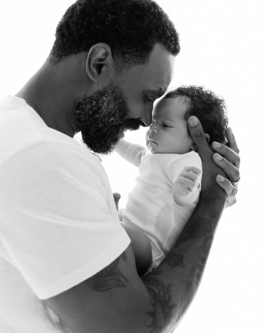 Black and white photo of a tattooed father tenderly holding his newborn baby, touching foreheads in a loving moment.