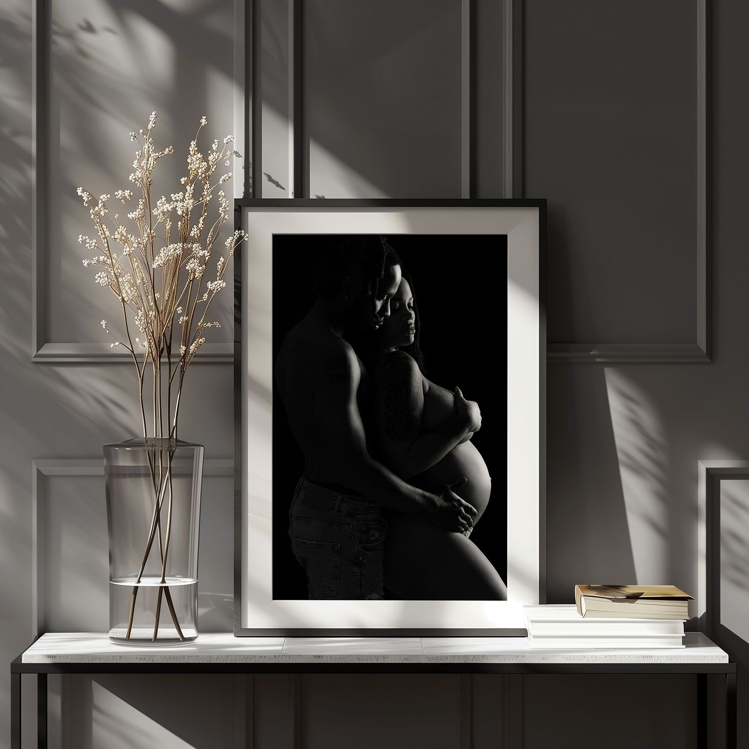 Artistic black and white maternity photograph displayed in frame on white shelf with dried flowers in vase.