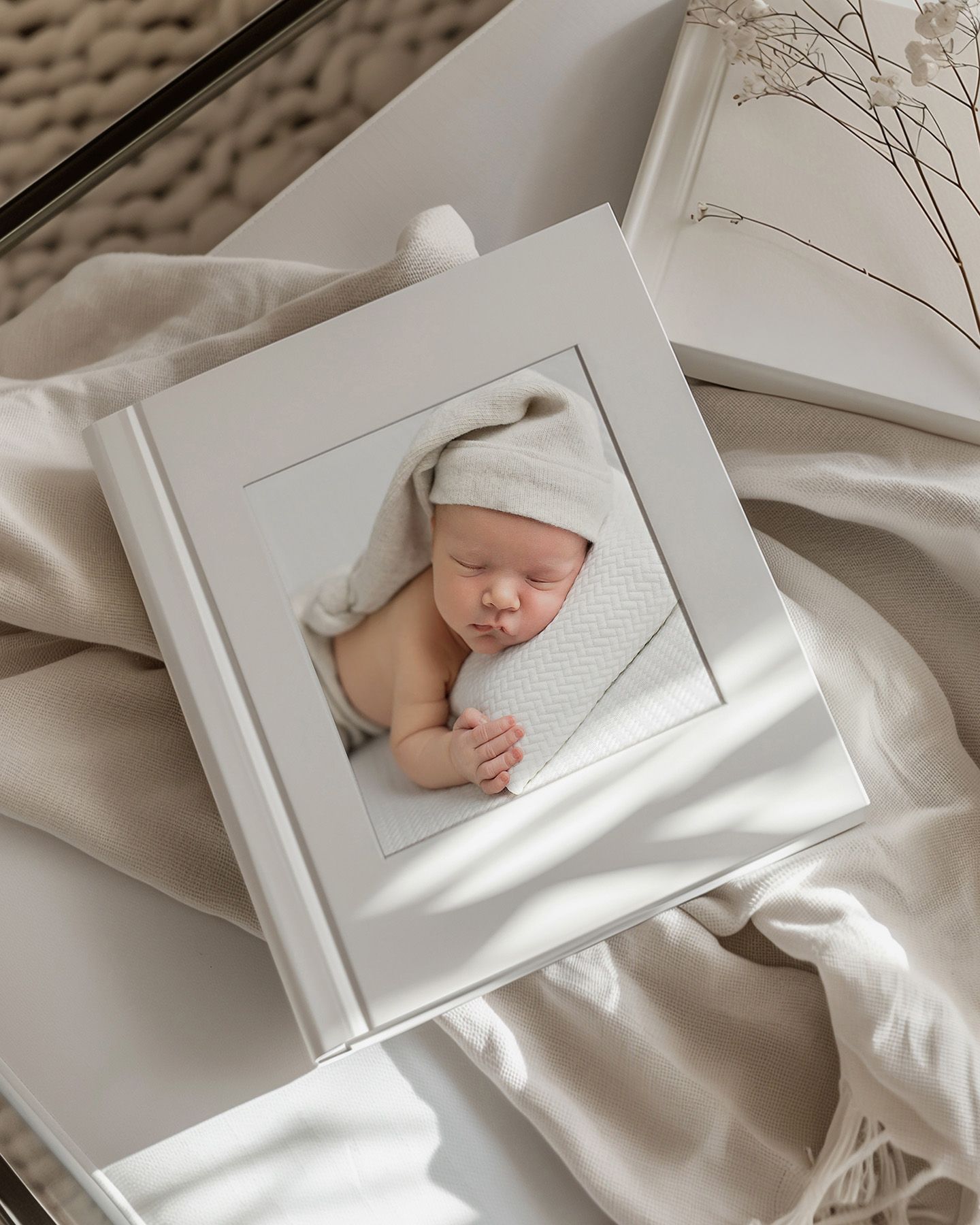 White photo album with a sleeping newborn baby wearing a knit hat, displayed on soft cream fabric.