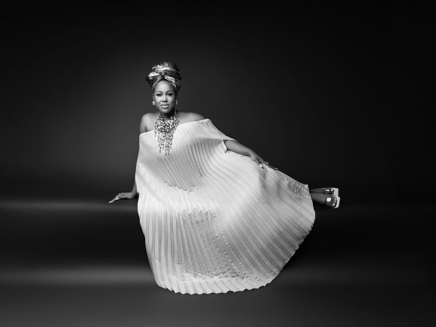 Black and white photo of a woman in a flowing pleated white gown and head wrap, posing elegantly.