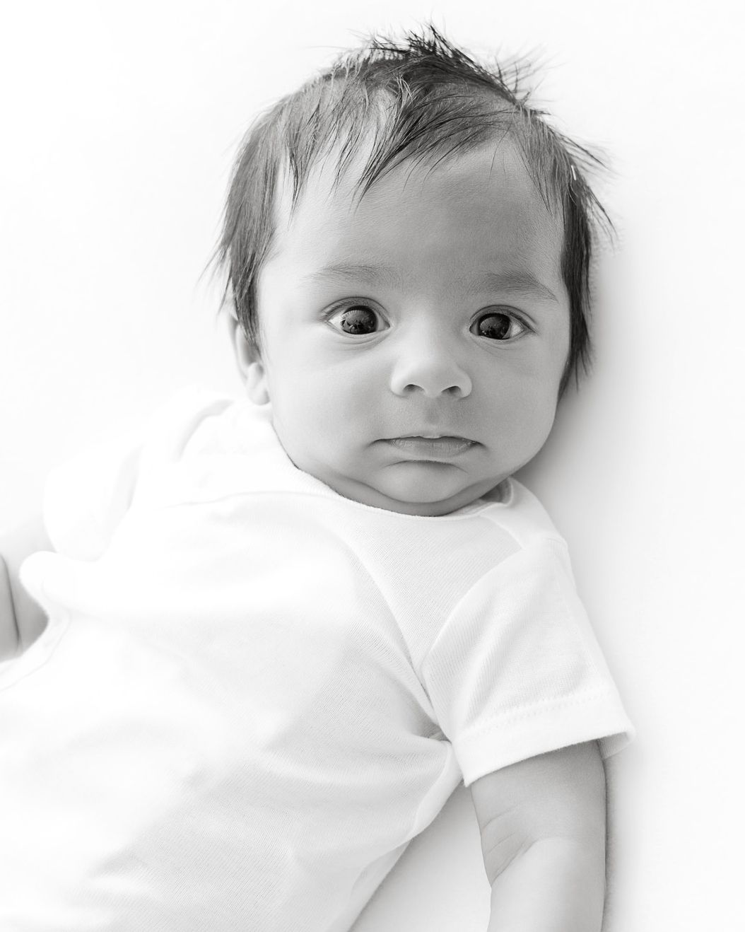 Black and white portrait of a newborn baby with dark hair wearing a white onesie, looking at the camera.