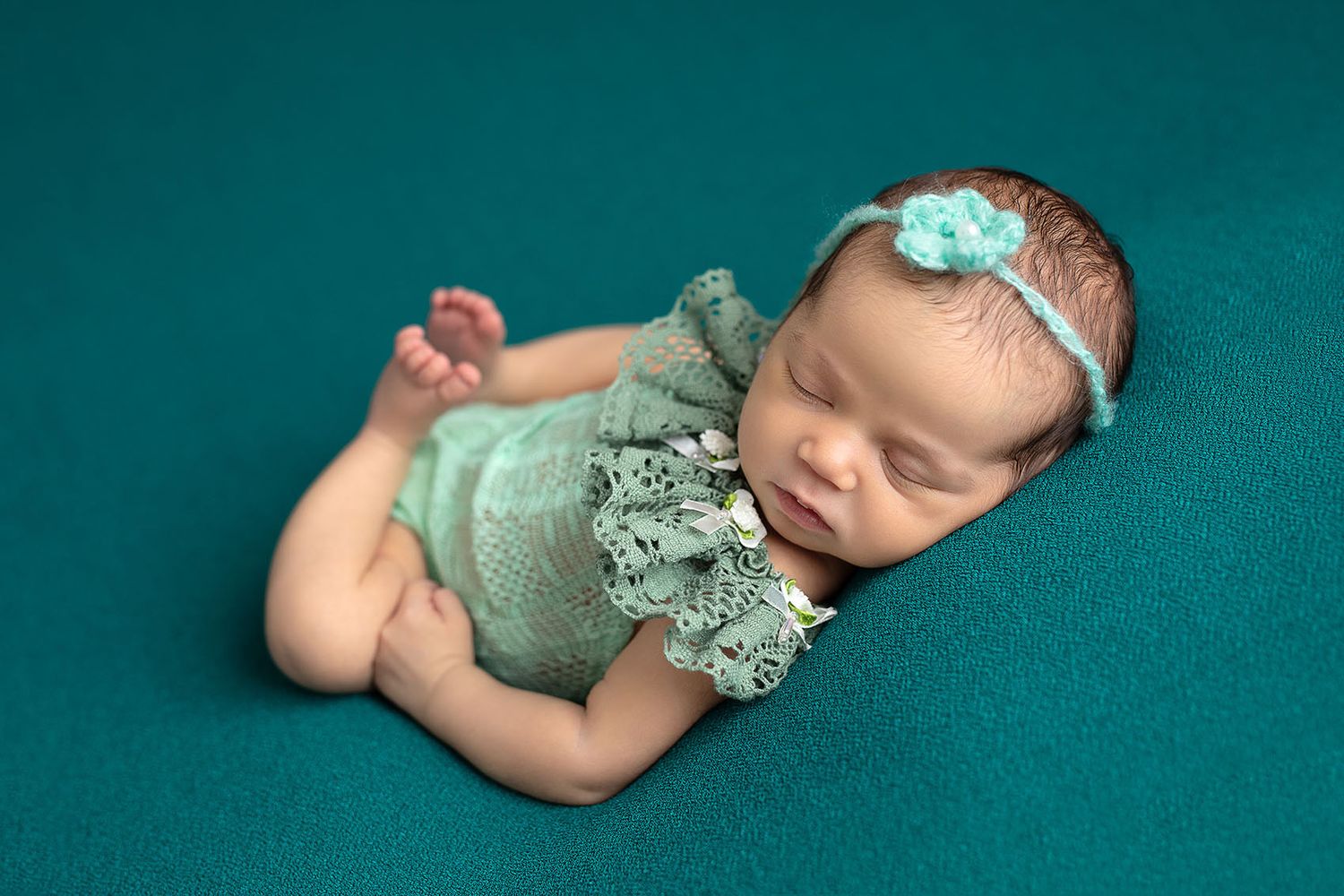 Newborn baby girl sleeping peacefully in teal lace romper and floral headband on teal backdrop.