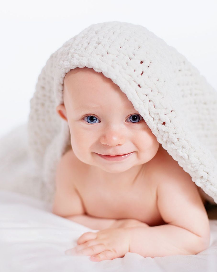 Adorable smiling baby with blue eyes lying under a white knit blanket on a soft white background.