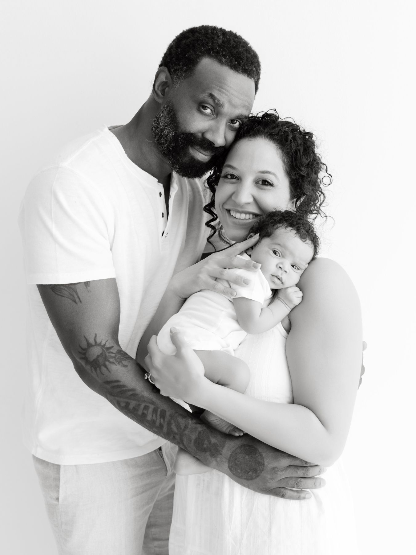 Black and white portrait of smiling couple in white outfits holding their newborn baby against white background.