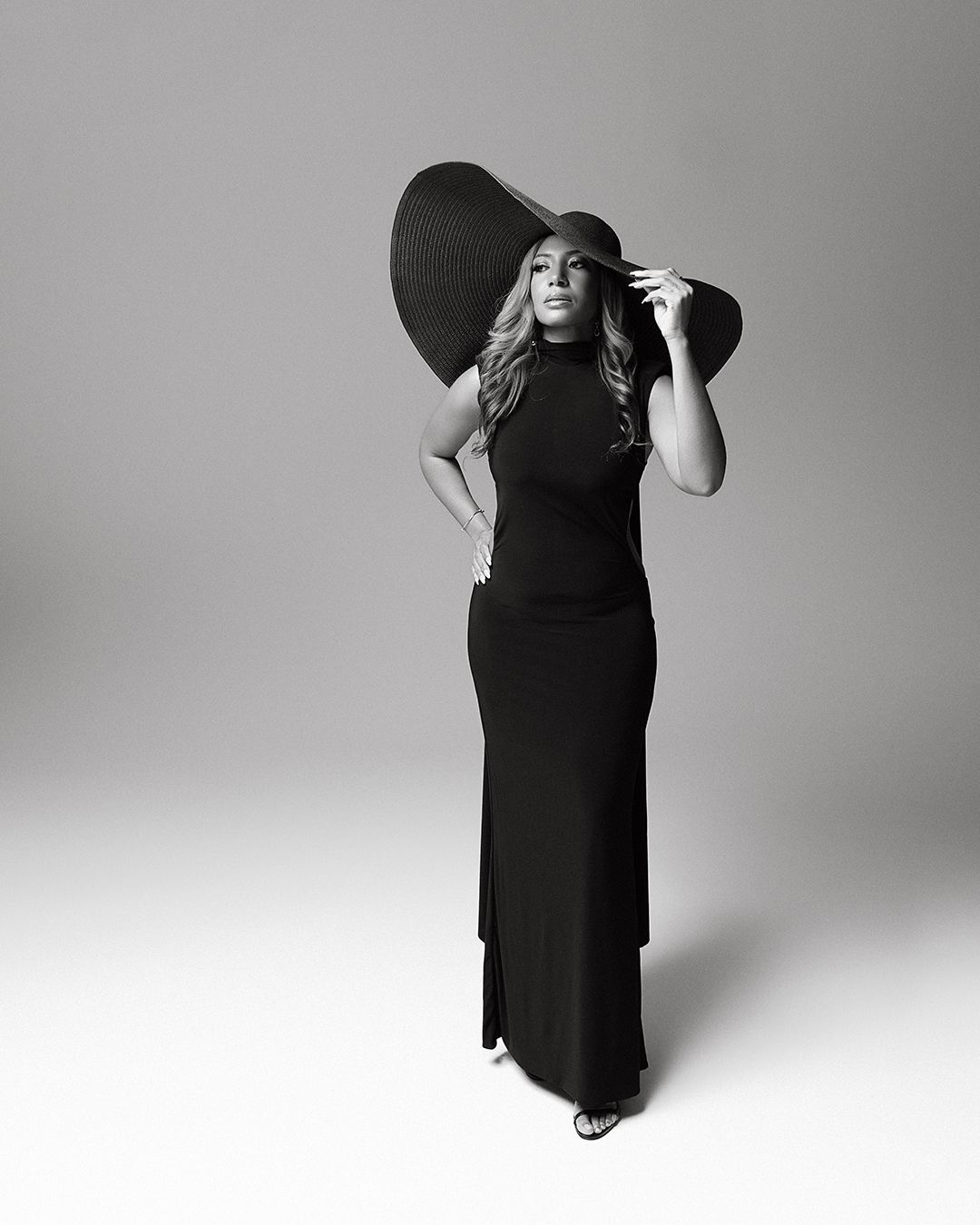 Black and white photo of a woman in a sleek black maxi dress and wide-brim hat posing elegantly.