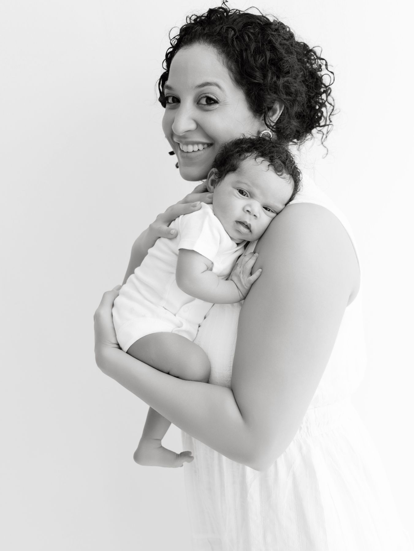 Black and white photo of a smiling mother with curly hair holding her newborn baby in a white onesie.