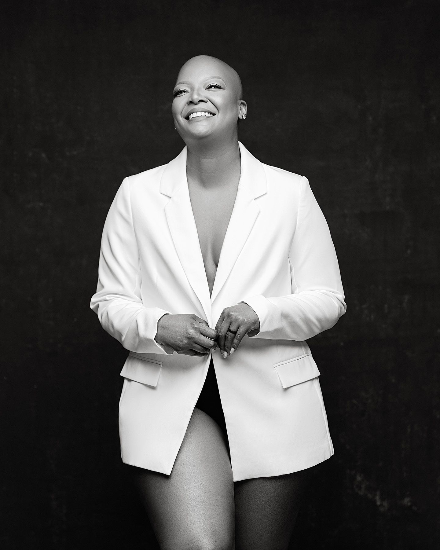 Black and white portrait of a smiling bald woman in a white blazer, posing confidently against a dark background.
