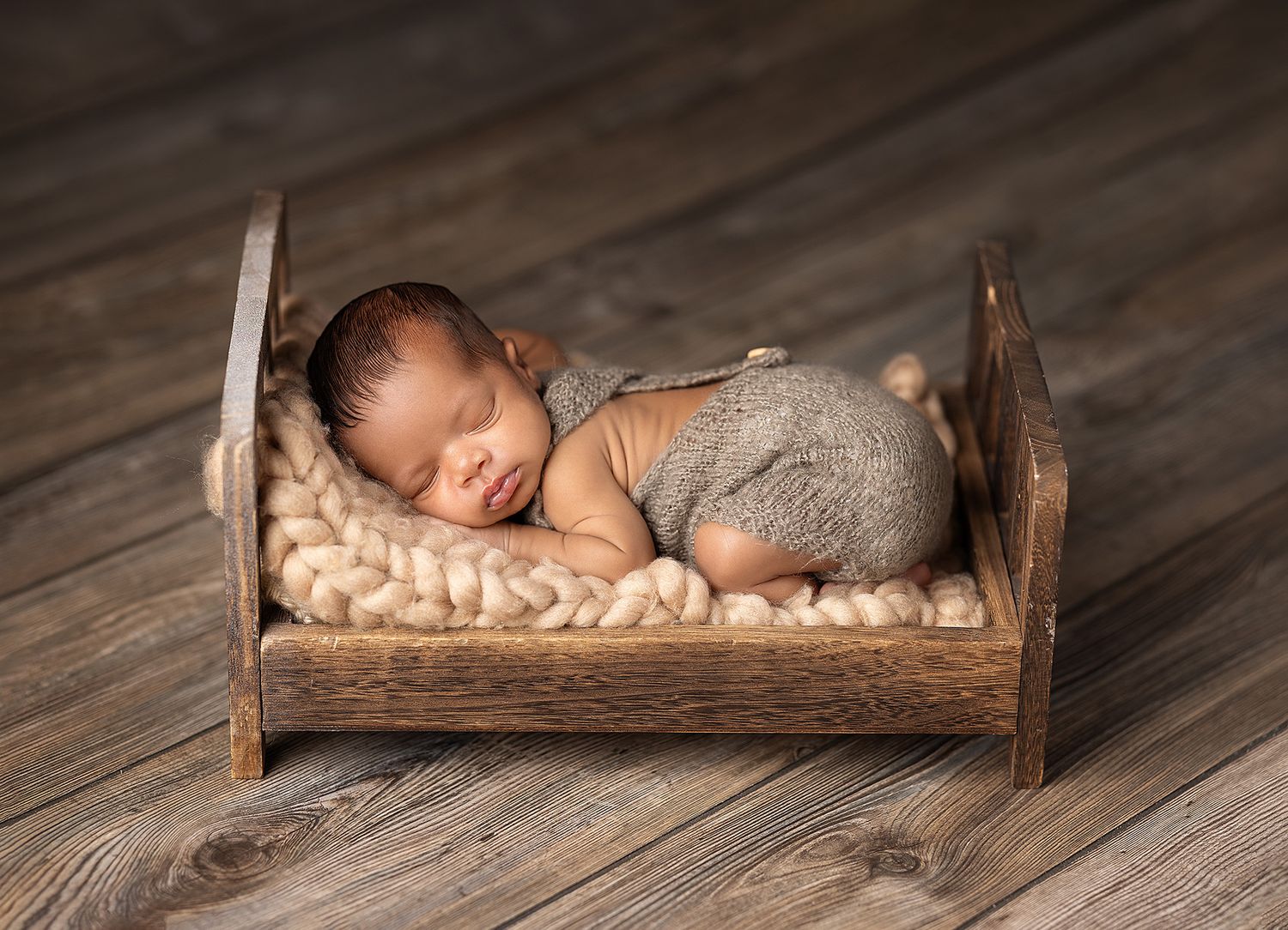 Newborn baby sleeping peacefully in a rustic wooden miniature bed with cream knit blanket, wearing a brown romper.