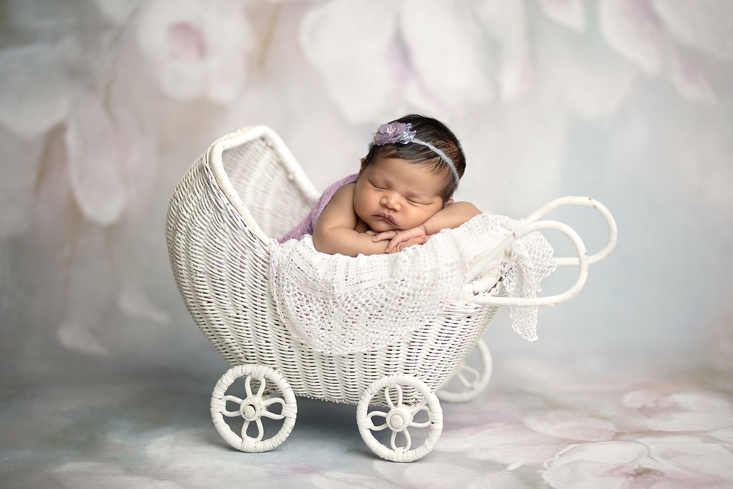 Newborn baby girl sleeping peacefully in a white wicker pram with a purple headband on a floral backdrop.
