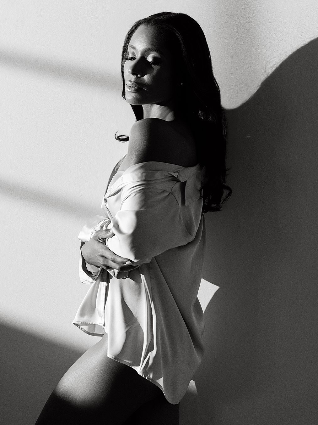 Black and white portrait of a woman in an off-shoulder top leaning against a wall in dramatic light.