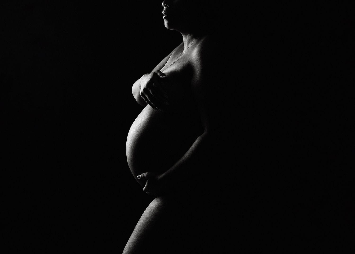Black and white silhouette of a pregnant woman cradling her baby bump against a dark background.