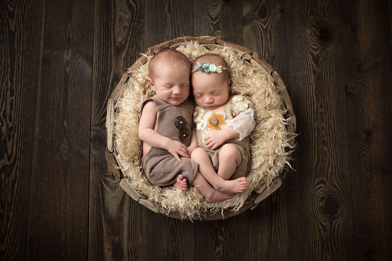 Two sleeping newborn twins curled together in a rustic basket with fluffy fur lining, one holding a teddy bear.