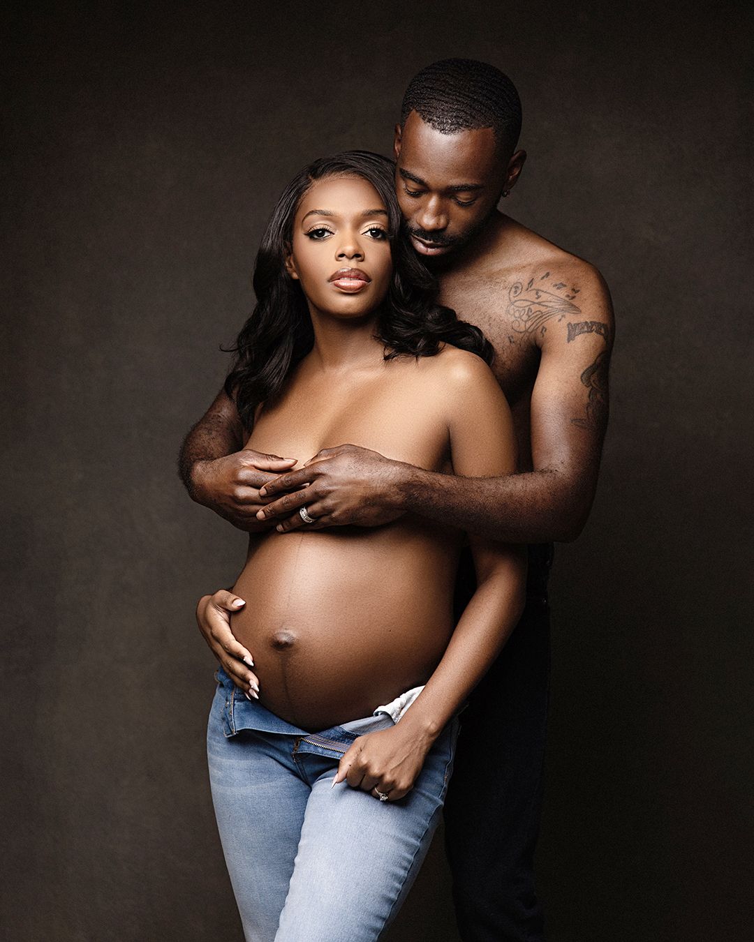 A couple poses for a maternity photo shoot, with the shirtless man embracing the pregnant woman from behind.