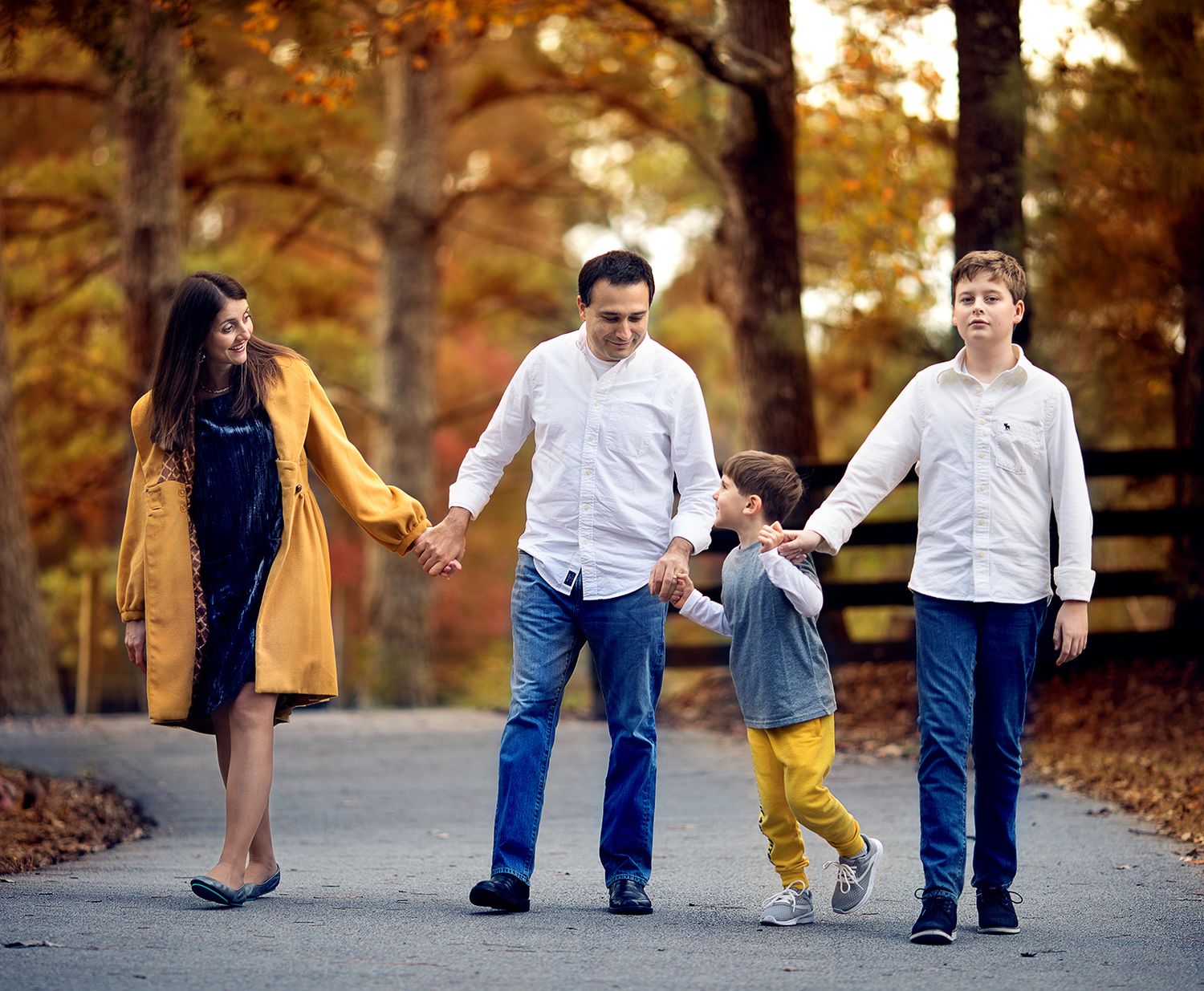 A family of four holds hands while walking together on a path surrounded by autumn trees with golden foliage.