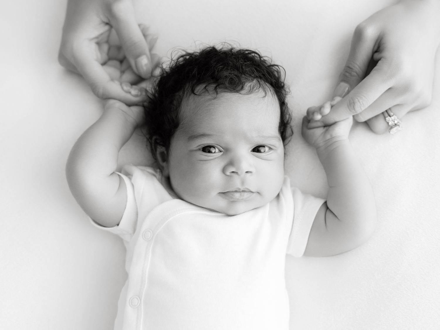 Black and white newborn photo of a alert baby in white onesie held by parents hands on white background.