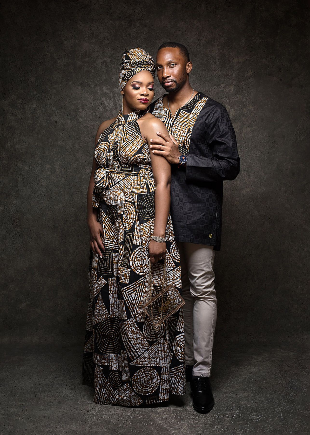 A couple in coordinating African-inspired outfits pose together against a dark textured backdrop in a studio portrait.