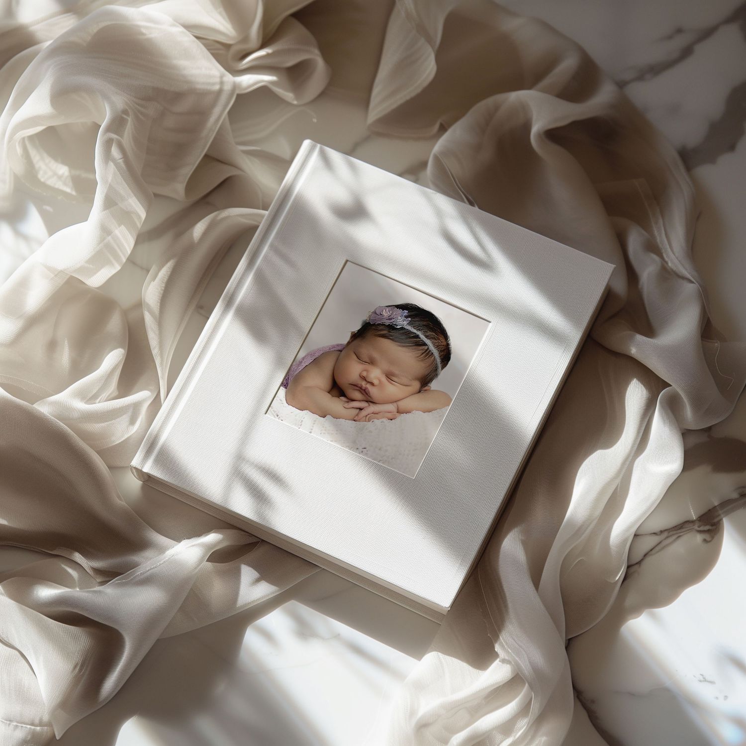 White hardcover baby photo album with newborn portrait, placed on soft ivory silk fabric in natural light.