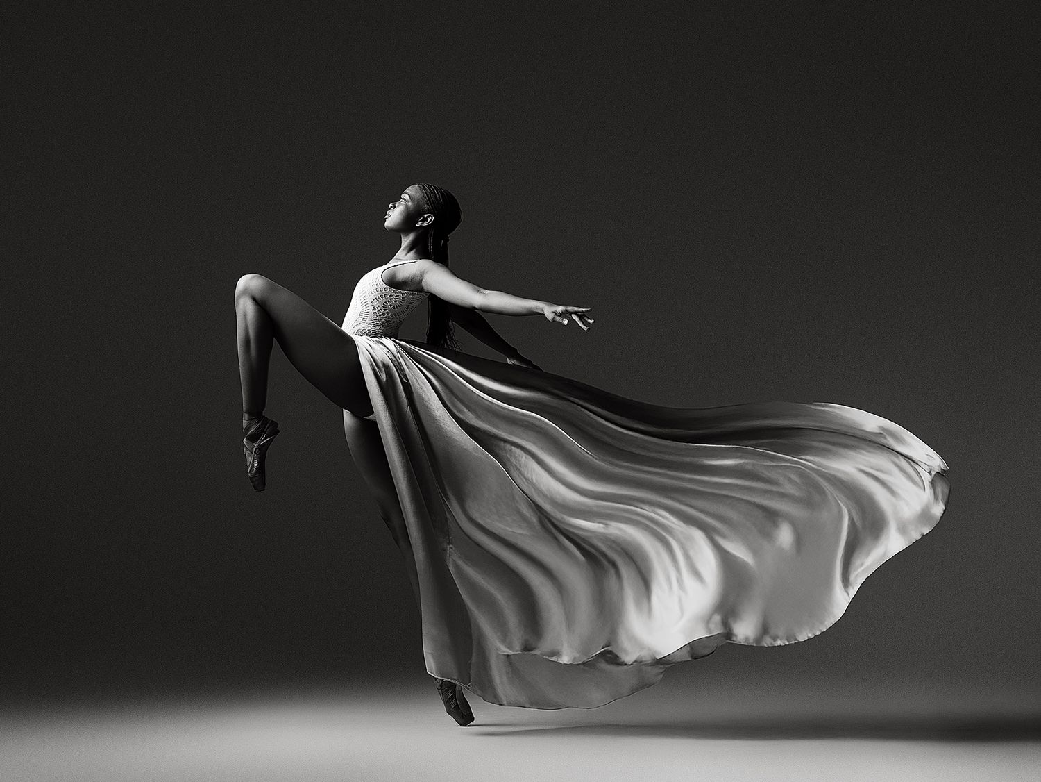 A graceful dancer in flowing white dress leaps through the air against dark background in dramatic black and white.